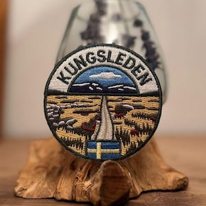 Op de afbeelding: Geborduurde ronde patch met de tekst "KUNGSLEDEN". Het ontwerp toont een landschap met bergen, een pad en de Zweedse vlag. De patch wordt gepresenteerd op een stuk hout.