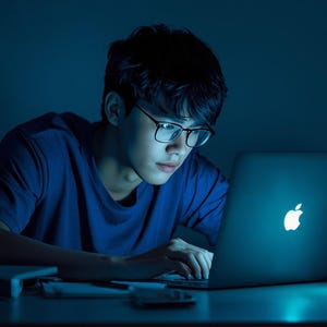 May include: A person wearing glasses and a blue t-shirt is focused on a laptop with the Apple logo illuminated. The scene is bathed in blue light, with stacks of books visible on the desk, suggesting a study or work environment.