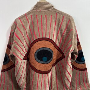 May include: Tan jacket with large embroidered eye designs on the back and sleeves. The eyes are in shades of brown, orange, and black, with red radiating lines. The jacket is hanging on a metal hanger.