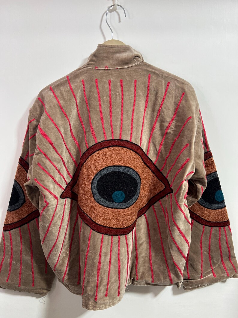 May include: Tan jacket with large embroidered eye designs on the back and sleeves. The eyes are in shades of brown, orange, and black, with red radiating lines. The jacket is hanging on a metal hanger.