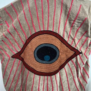 May include: A tan jacket with a large embroidered eye design on the back. The eye is brown, black, blue, and orange, with red embroidered rays emanating from it. The jacket has a collar and a soft, velvety texture.