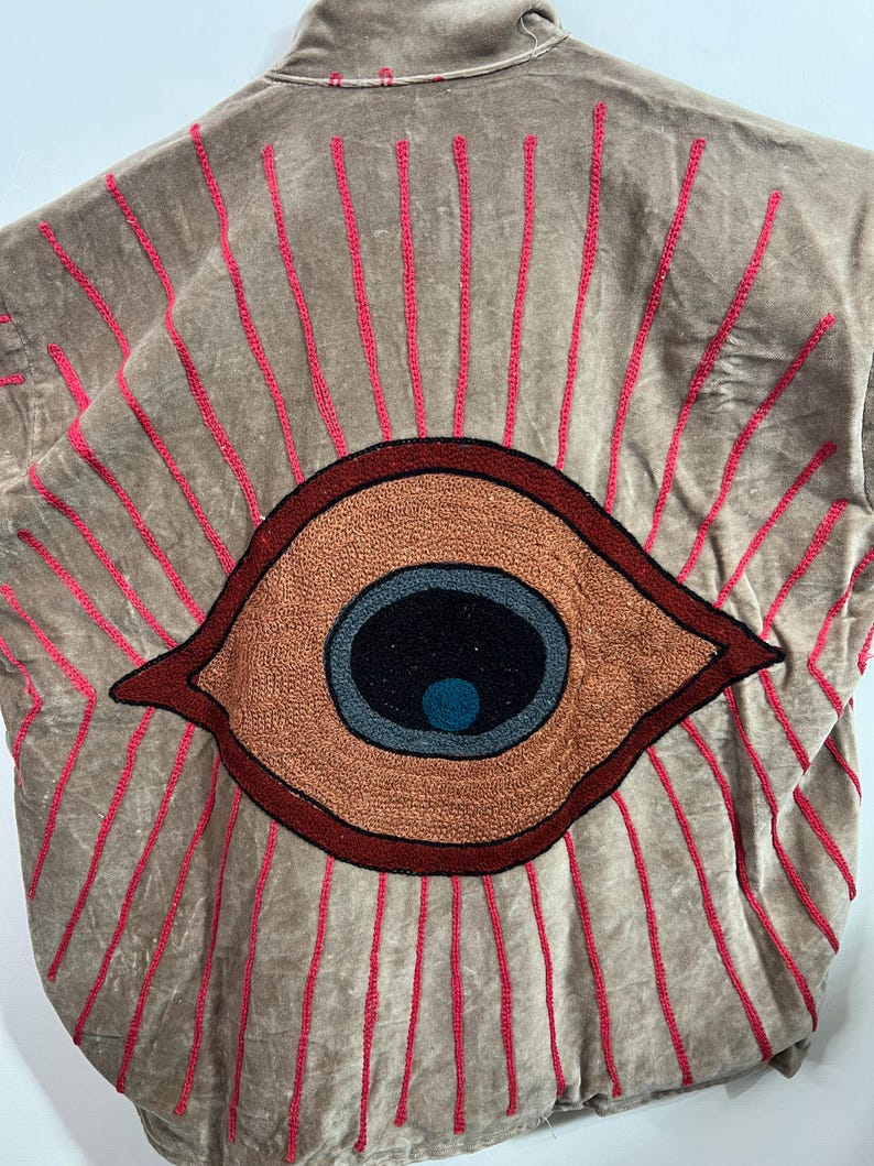 May include: A tan jacket with a large embroidered eye design on the back. The eye is brown, black, blue, and orange, with red embroidered rays emanating from it. The jacket has a collar and a soft, velvety texture.
