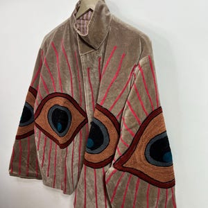 May include: A tan velvet jacket with red vertical stripes and large embroidered eye designs on the sleeves. The jacket has a high collar and is hanging on a wooden hanger.