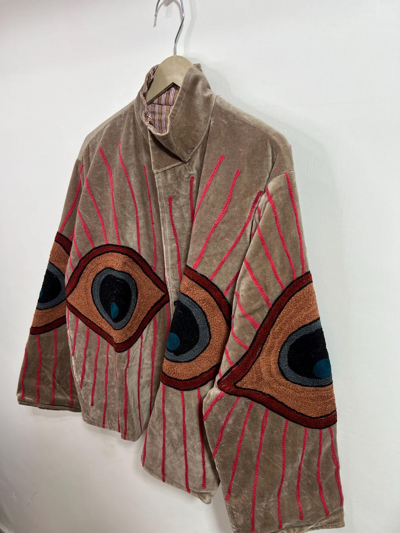 May include: A tan velvet jacket with red vertical stripes and large embroidered eye designs on the sleeves. The jacket has a high collar and is hanging on a wooden hanger.