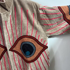 May include: A tan velvet jacket with embroidered details. The jacket features large eye designs in shades of brown, orange, and blue, with red vertical lines. The sleeves have similar eye designs and red horizontal lines.