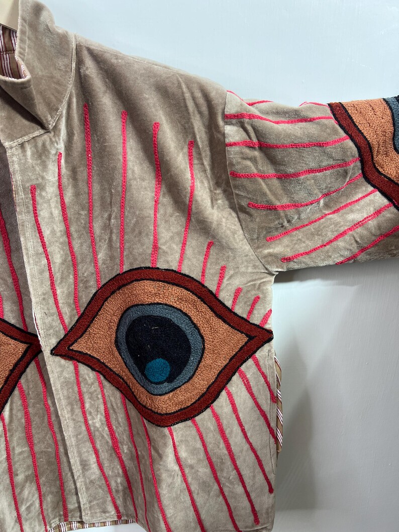 May include: A tan velvet jacket with embroidered details. The jacket features large eye designs in shades of brown, orange, and blue, with red vertical lines. The sleeves have similar eye designs and red horizontal lines.