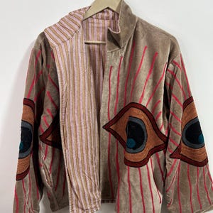 May include: A tan velvet jacket with red vertical stripes and embroidered eye designs on the sleeves. The jacket has a striped interior and is hanging on a wooden hanger.