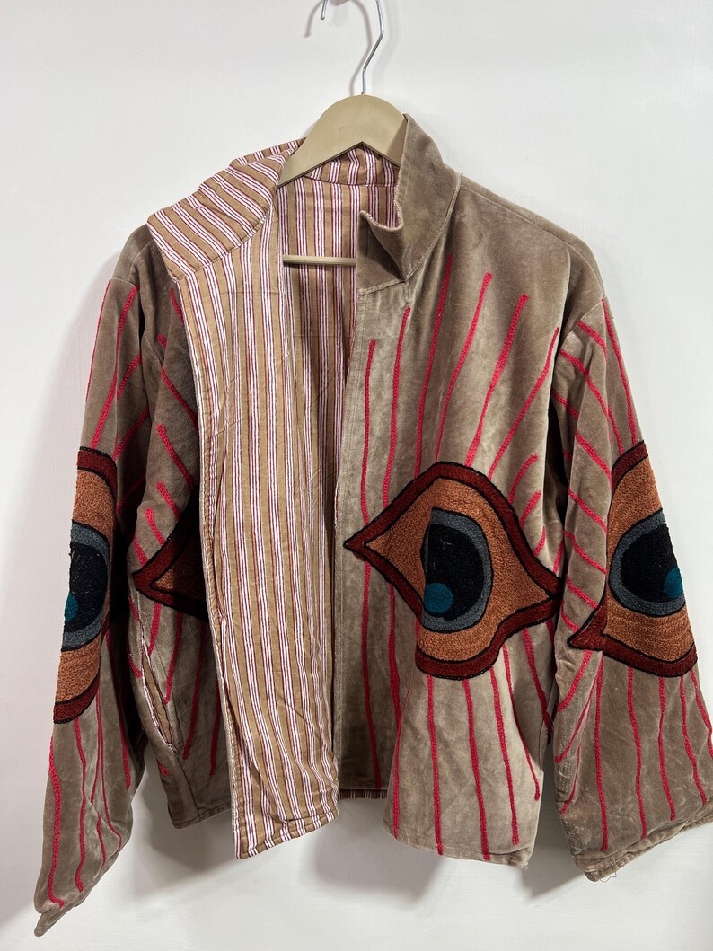 May include: A tan velvet jacket with red vertical stripes and embroidered eye designs on the sleeves. The jacket has a striped interior and is hanging on a wooden hanger.