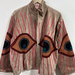 May include: A tan velvet jacket with embroidered red vertical lines and large eye designs on the sleeves. The eyes are brown, black, and teal. The jacket has a high collar and is hanging on a metal hanger.
