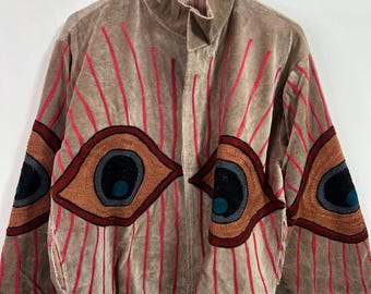 Könnte beinhalten: Eine beige Samtjacke mit gestickten roten vertikalen Linien und großen Augendesigns auf den Ärmeln. Die Augen sind braun, schwarz und türkis. Die Jacke hat einen hohen Kragen und hängt an einem Metallbügel.