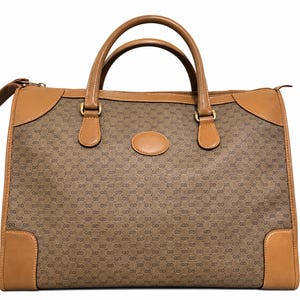 May include: A structured handbag featuring a brown canvas body with a repeating dark brown logo pattern. The bag includes tan leather trim, two top handles, a zipper closure, and a central oval leather patch on the front exterior panel.