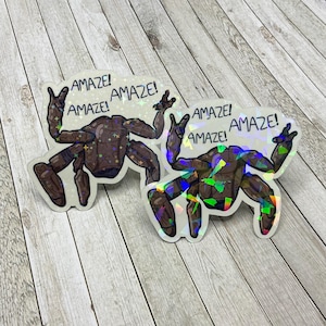 May include: Two die-cut stickers featuring a brown, multi-legged creature with a segmented body. The creature has raised arms and the text "AMAZE!" printed three times above it. The stickers have a holographic finish with star and shard patterns.