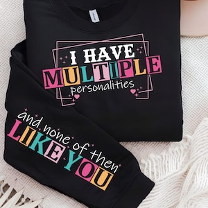 Peut inclure: Sweat-shirt noir avec l'inscription "I HAVE MULTIPLE personalities" dans une police de caractères colorée et en blocs. La manche affiche "and none of then LIKE YOU" dans un style similaire. Le design est sur un fond neutre.