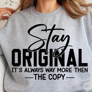 May include: A light gray sweatshirt with the words "Stay Original" in black script and block letters. Below, the text reads "It's Always Way More Then - The Copy -" in black. The sweatshirt is a casual style.