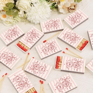 Op de afbeelding: Luciferdoosjes met een roze en wit gestreept ontwerp. Elke doos heeft een rood hartvormig ontwerp met de letters "R" en "Y". De luciferkoppen zijn rood. Een paar lucifers liggen verspreid rond de dozen. Het thema is waarschijnlijk gerelateerd aan bruiloften of romantiek.