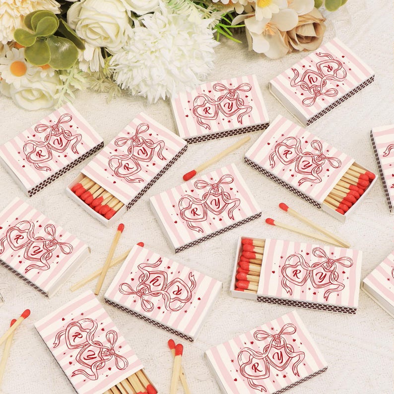 Op de afbeelding: Luciferdoosjes met een roze en wit gestreept ontwerp. Elke doos heeft een rood hartvormig ontwerp met de letters "R" en "Y". De luciferkoppen zijn rood. Een paar lucifers liggen verspreid rond de dozen. Het thema is waarschijnlijk gerelateerd aan bruiloften of romantiek.