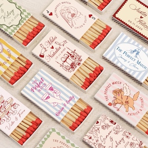 Op de afbeelding: Een verzameling decoratieve lucifersdoosjes met rode lucifers. Elke doos heeft unieke ontwerpen en tekst, waaronder namen en data, met thema's van bruiloften en feesten. De dozen zijn wit, met verschillende kleuren en illustraties.