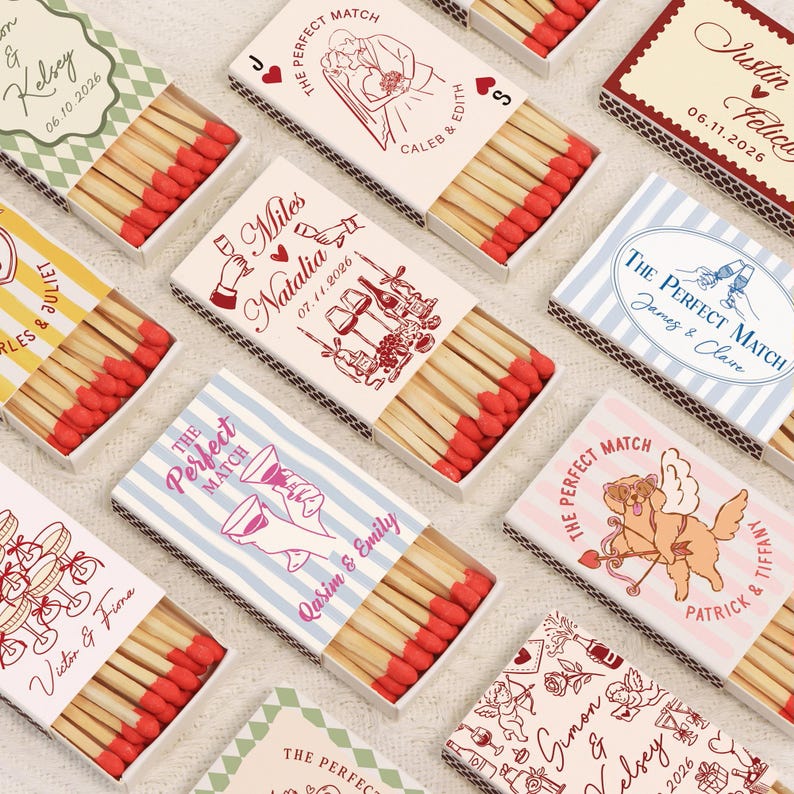 Op de afbeelding: Een verzameling decoratieve lucifersdoosjes met rode lucifers. Elke doos heeft unieke ontwerpen en tekst, waaronder namen en data, met thema's van bruiloften en feesten. De dozen zijn wit, met verschillende kleuren en illustraties.