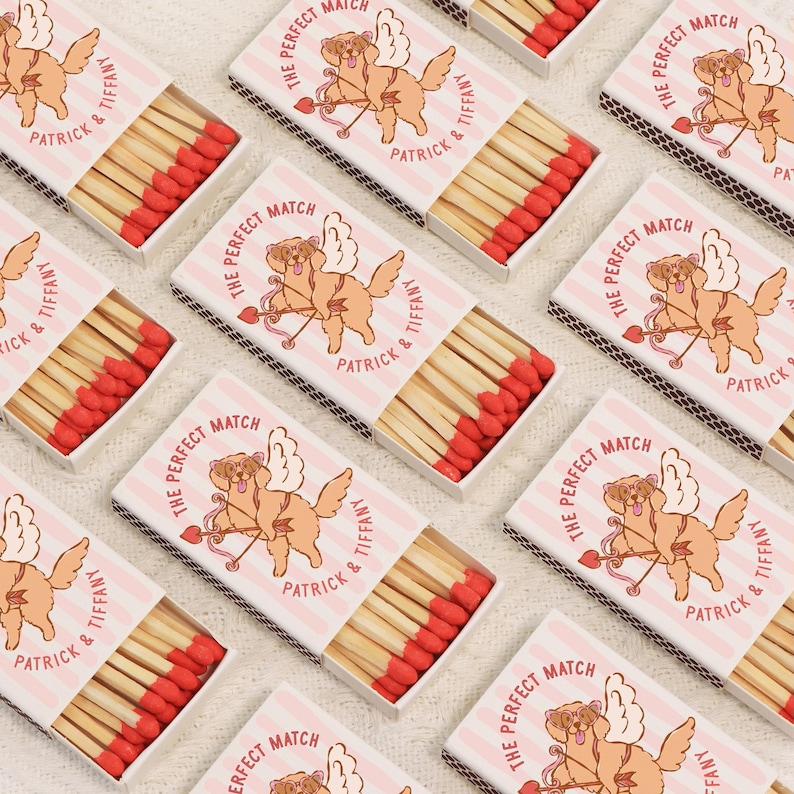 Op de afbeelding: Meerdere lucifersdozen met rode koppen zijn gerangschikt op een wit oppervlak. Elke doos heeft een roze en wit gestreept ontwerp met een cartoonhond met vleugels en pijl en boog. De tekst "THE PERFECT MATCH" en "PATRICK & TIFFANY" staan op de dozen.