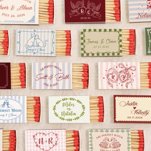 Op de afbeelding: Een assortiment gepersonaliseerde lucifersdoosjes met diverse ontwerpen en tekst. Sommige doosjes tonen namen en data, terwijl andere illustraties van katten, harten en bloemmotieven hebben. De luciferkoppen zijn rood.