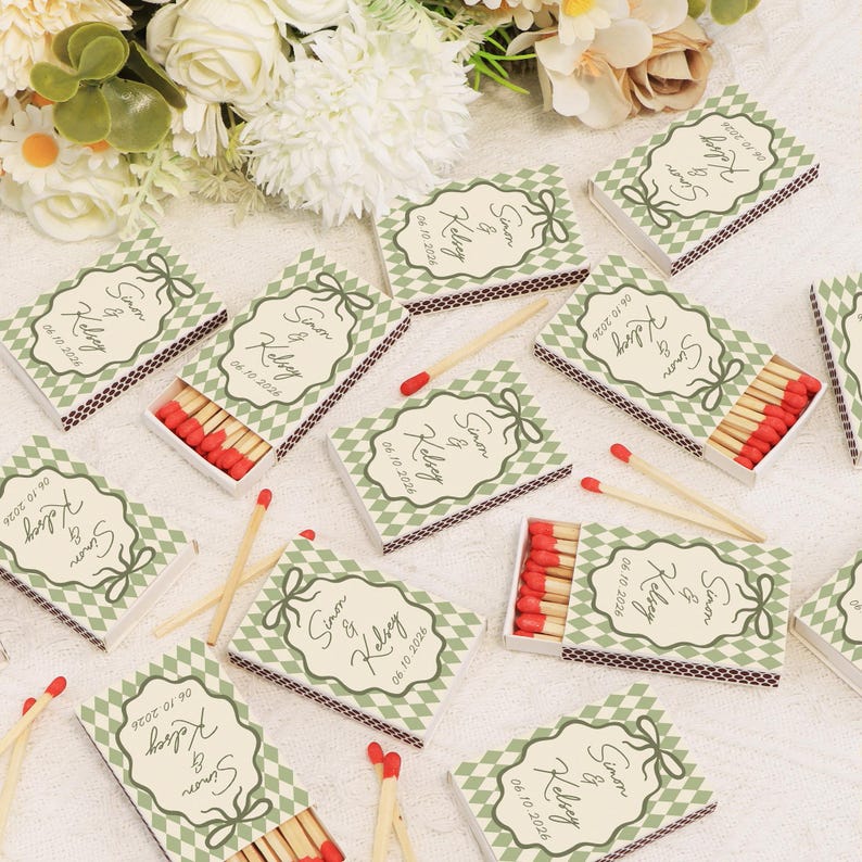 Op de afbeelding: Luciferdoosjes met een groen en wit ruitpatroon en een cr&egrave;mekleurig label met de namen "Simon & Kelsey" en de datum "06.10.2026". De luciferkoppen zijn rood.