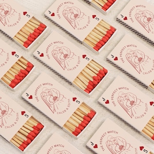 Op de afbeelding: Verschillende lucifersdozen met rode koppen zijn gerangschikt op een wit oppervlak. Elke doos heeft een rode illustratie van een stel en de tekst "The Perfect Match", samen met namen en kaartsymbolen.