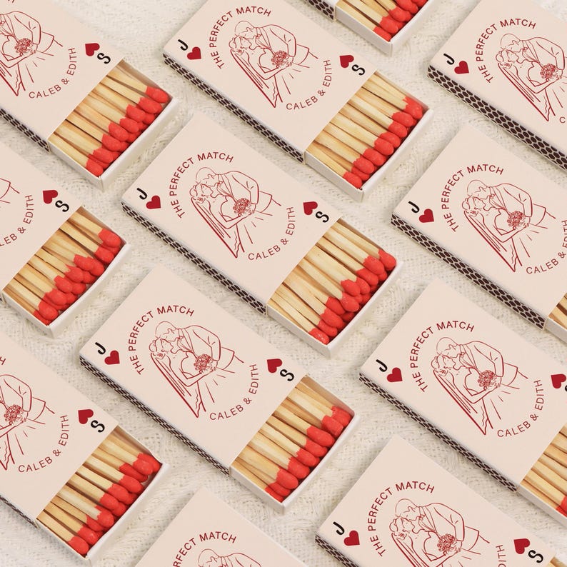 Op de afbeelding: Verschillende lucifersdozen met rode koppen zijn gerangschikt op een wit oppervlak. Elke doos heeft een rode illustratie van een stel en de tekst "The Perfect Match", samen met namen en kaartsymbolen.