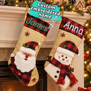Puede incluir: Dos calcetines navideños de arpillera color canela con puños de cuadros rojos y negros. Uno tiene el nombre bordado James en verde, el otro Anna en blanco. Ambos incluyen aplicaciones de Papá Noel o muñeco de nieve y copos de nieve rojos.
