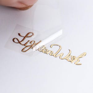 May include: A gold-colored metallic decal featuring the cursive script text "LightenWish" attached to a clear adhesive transfer film. The decal is partially peeled back from the backing, revealing the shiny, reflective surface of the lettering.