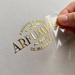 May include: A gold foil sticker featuring the text ANTONIO & GINO ARFUMERIA DI MARRAZZO in a curved layout with decorative flourishes. The sticker is partially peeled back from its clear backing, revealing the metallic finish and adhesive design.
