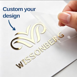 May include: A gold metallic decal featuring a stylised emblem and the text WISSONBERG being applied to a white surface with a clear transfer film. The text Custom your design is printed in blue with a blue arrow pointing to the gold logo.