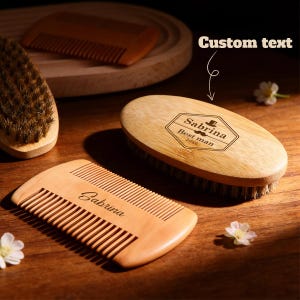 Puede incluir: Set de cuidado de barba de madera con un cepillo y dos peines. El cepillo tiene el texto "Sabrina Best man 2026" y un gráfico de bigote. Un peine está grabado con "Sabrina". El set está sobre una superficie de madera con pequeñas flores blancas.