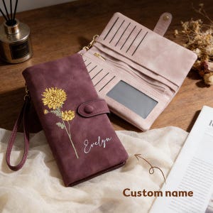 Puede incluir: Se muestran dos carteras, una burdeos y otra rosa claro. La cartera burdeos presenta un diseño floral amarillo y el nombre "Evelyn" en escritura blanca. La cartera rosa claro está abierta, revelando ranuras para tarjetas y una ventana de identificación transparente.