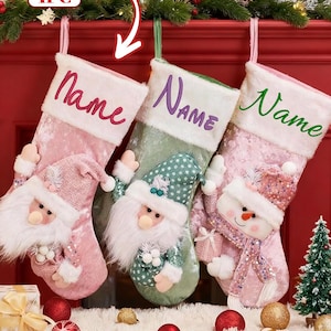 Op de afbeelding: Drie kerstsokken met figuren van de kerstman en een sneeuwpop. De sokken zijn roze en groen, met witte randen en het woord "Name" geborduurd. Een kleine kerstboom en ornamenten staan op de voorgrond.