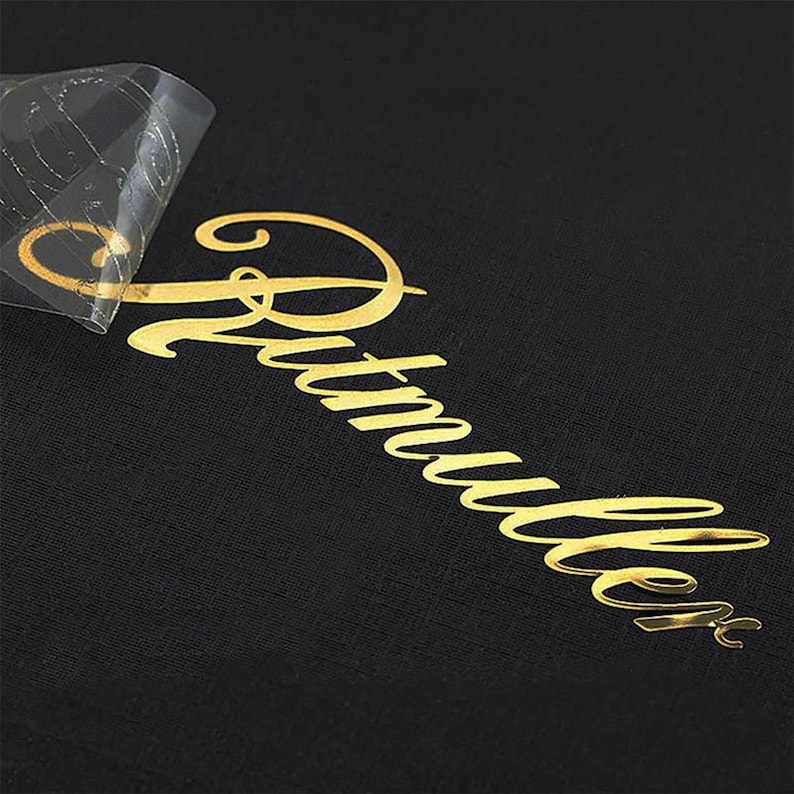 May include: A black fabric surface featuring a metallic gold heat transfer vinyl decal with the script text "Rutmullet". A portion of the clear adhesive transfer film is partially peeled back from the top left corner of the gold lettering.