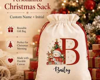 Puede incluir: Un saco de Navidad de lona color crema con cordones rojos. La parte frontal presenta una letra B grande en rojo, un gráfico de un muñeco de nieve y un reno junto a un árbol decorado, y el nombre Bailey en letras cursivas negras.