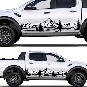 May include: White pickup truck with black mountain and tree decals on the side. The decals feature a silhouette of a mountain range and evergreen trees, creating a nature-themed design. The truck has black wheels and tires.