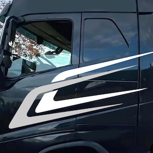 May include: Black semi-truck cab with white and gray decorative decals. The decals are a stylized design that curves along the side of the cab. The truck has tinted windows and a black roof. The image is taken outdoors with a cloudy sky in the background.