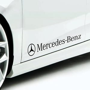 Pode incluir: Autocolante preto com o logótipo e texto Mercedes-Benz na lateral de um carro branco. O logótipo apresenta uma estrela de três pontas dentro de um círculo. O texto está em uma fonte sem serifa.