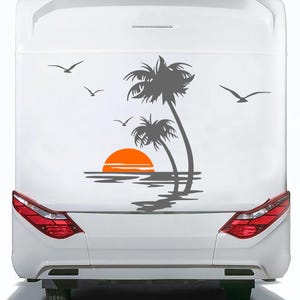 Puede incluir: Parte trasera de un vehículo blanco con una calcomanía de palmera y puesta de sol gris. La calcomanía presenta dos palmeras, un sol naranja y un reflejo en el agua. También se representan varios pájaros volando.