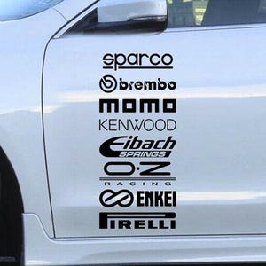Könnte beinhalten: Weißes Auto mit schwarzen Rennaufklebern. Die Aufkleber enthalten die Marken Sparco, Brembo, MOMO, Kenwood, Eibach Springs, O.Z Racing, Enkei und Pirelli. Das Auto hat ein orangefarbenes Rad.