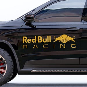 Könnte beinhalten: Ein schwarzes Fahrzeug mit goldfarbenen Buchstaben und Grafiken. Der Text "Red Bull RACING" ist an der Seite zu sehen. Oberhalb des Textes befindet sich eine Grafik von zwei sich gegenüberstehenden Bullen mit einer Sonne im Hintergrund. Das Fahrzeug hat schwarze Reifen und einen schwarzen Dachträger.