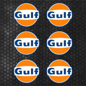 Puede incluir: Seis pegatinas circulares con el logotipo de Gulf Oil. Cada pegatina presenta un círculo naranja con un óvalo horizontal blanco y la palabra "Gulf" en azul. Están dispuestas sobre un fondo texturizado gris oscuro.