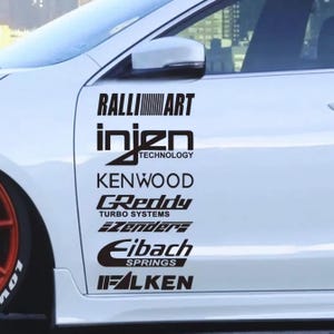 Op de afbeelding: Witte autodeur met zwarte stickers met verschillende merknamen, waaronder RALLIART, Injen Technology, KENWOOD, GReddy Turbo Systems en Eibach Springs. De auto heeft een oranje wiel.