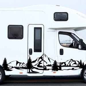May include: A white recreational vehicle with a black mountain and forest decal along the side. The decal features silhouettes of pine trees, mountains, and a lake. The RV has black windows and a door.