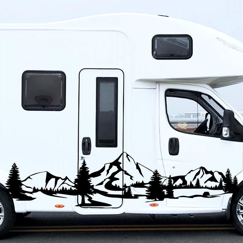 May include: A white recreational vehicle with a black mountain and forest decal along the side. The decal features silhouettes of pine trees, mountains, and a lake. The RV has black windows and a door.