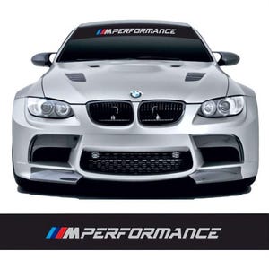 May include: Silver BMW car with a black windscreen banner displaying "IMPERFORMANCE" in white text with blue and red accents. The car features black grills and headlights. The image is a front view of the car.