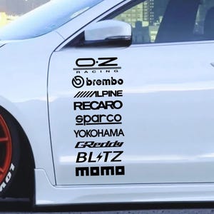 Pode incluir: Uma porta de carro branca com uma coluna vertical de autocolantes pretos de marcas automóveis. Os logótipos incluem O.Z Racing, Brembo, Alpine, Recaro, Sparco, Yokohama, GReddy, Blitz e Momo, dispostos numa única pilha no painel lateral.