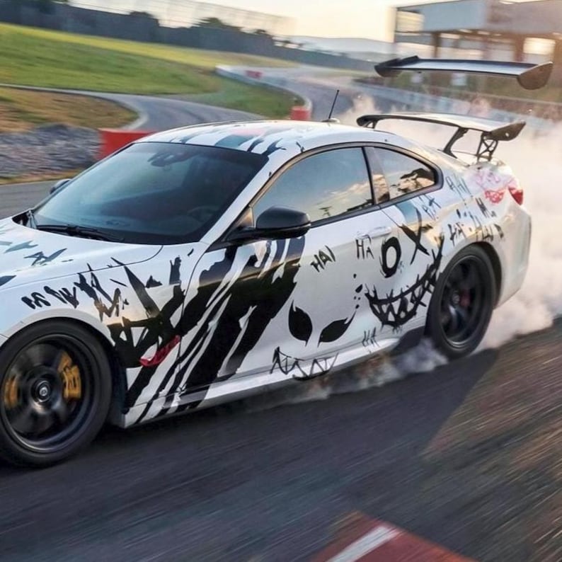 May include: A white sports car drifting on a track, emitting thick white smoke from its rear tires. The car features a black graphic design with sharp, jagged lines and a stylized face with a wide, toothy grin and the repeated text HA HA.