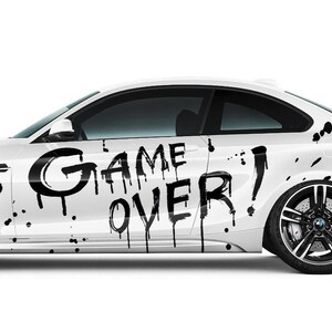 Peut inclure: Voiture blanche avec graffiti noir "GAME OVER" et éclaboussures de peinture noire. La voiture a des jantes noires et des vitres teintées. La voiture est de style coupé.